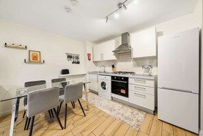 A modern kitchen with white cabinets, a stainless steel range hood, a built-in oven, and a refrigerator. There is a dining table with chairs and a rug on the floor. - 2 bed Nine Elms Apartment with Balcony: Easy Access to City & Underground (London)