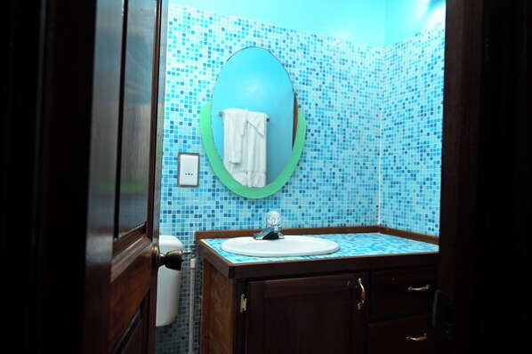 Une salle de bain avec un mur recouvert de mosaïques bleues, un miroir rond doté d’un cadre vert, un lavabo blanc et des meubles en bois.