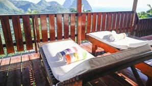 Una terraza de madera con dos sillones de descanso, cada uno con una toalla enrollada. La terraza ofrece vistas a un paisaje montañoso y al océano.