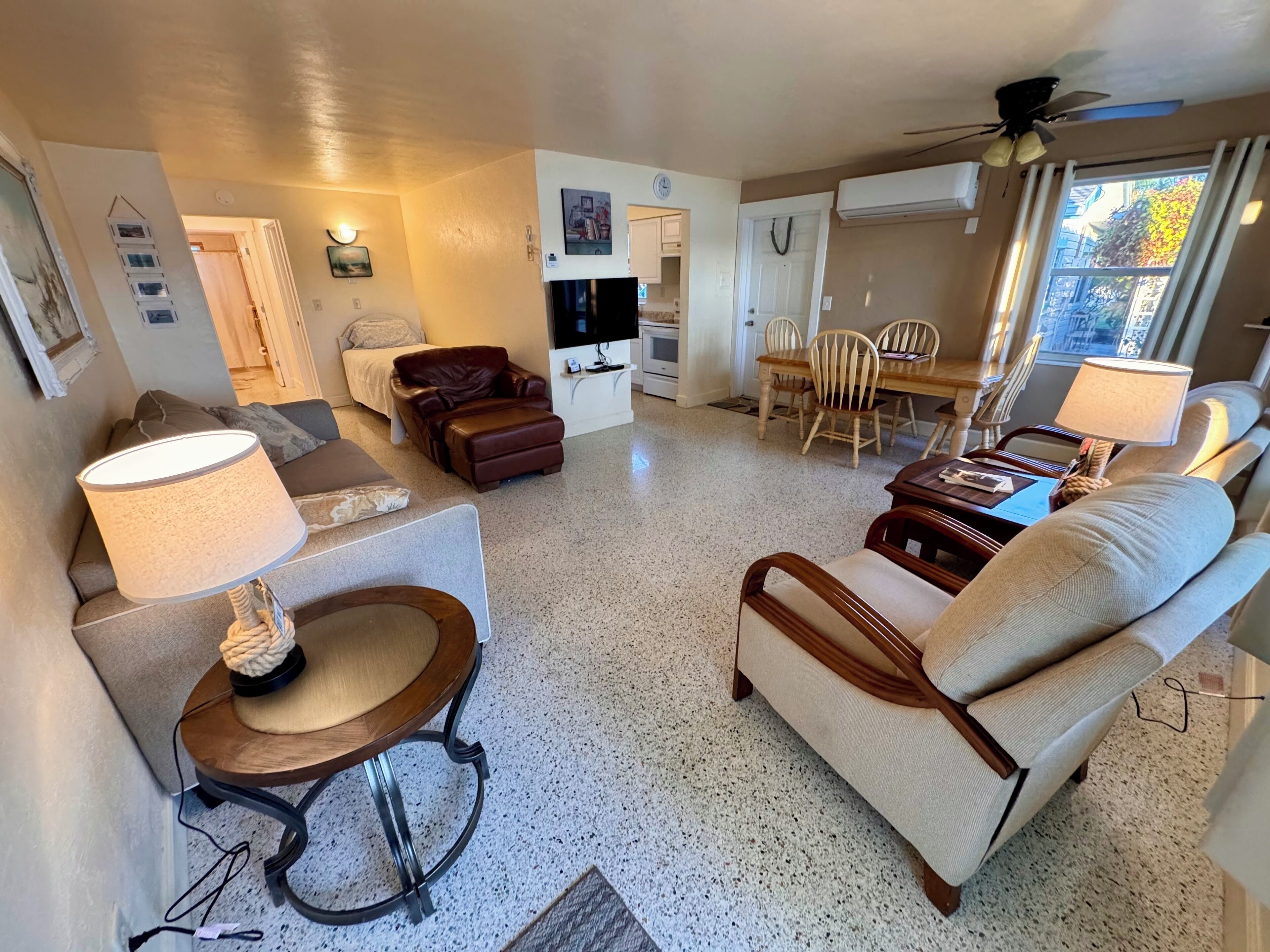 A living room with a sofa, armchair, and a dining table with chairs. There is a TV, a ceiling fan, and an air conditioning unit.