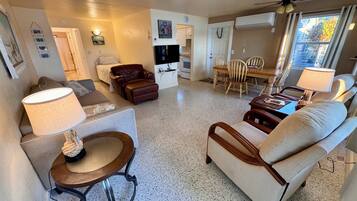 A living room with a sofa, armchair, and a dining table with chairs. There is a TV, a ceiling fan, and an air conditioning unit.