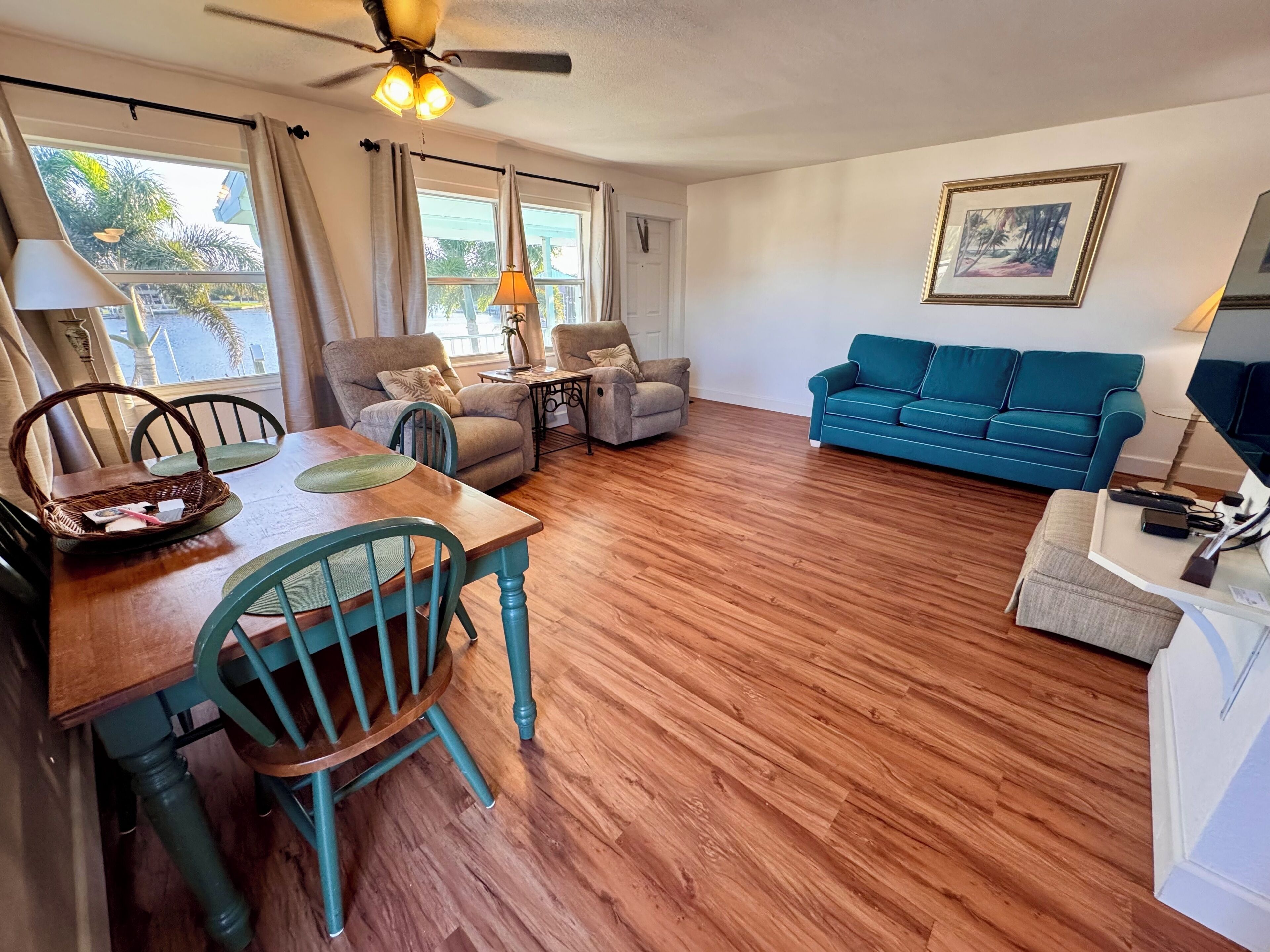 A living room with a wooden floor, a blue sofa, a dining table with chairs, and a ceiling fan.