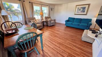 A living room with a wooden floor, a blue sofa, a dining table with chairs, and a ceiling fan.