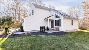 A modern two-story house with a sloped roof, a front porch with a bench, and a small garden area with shrubs and a tree. - Charming 3-bedroom house with AC in peaceful Tiverton (Tiverton)