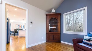 A spacious living room with hardwood flooring, a dark brown leather sofa, a wooden cabinet, and a large window with a view of trees. - Charming 3-bedroom house with AC in peaceful Tiverton (Tiverton)