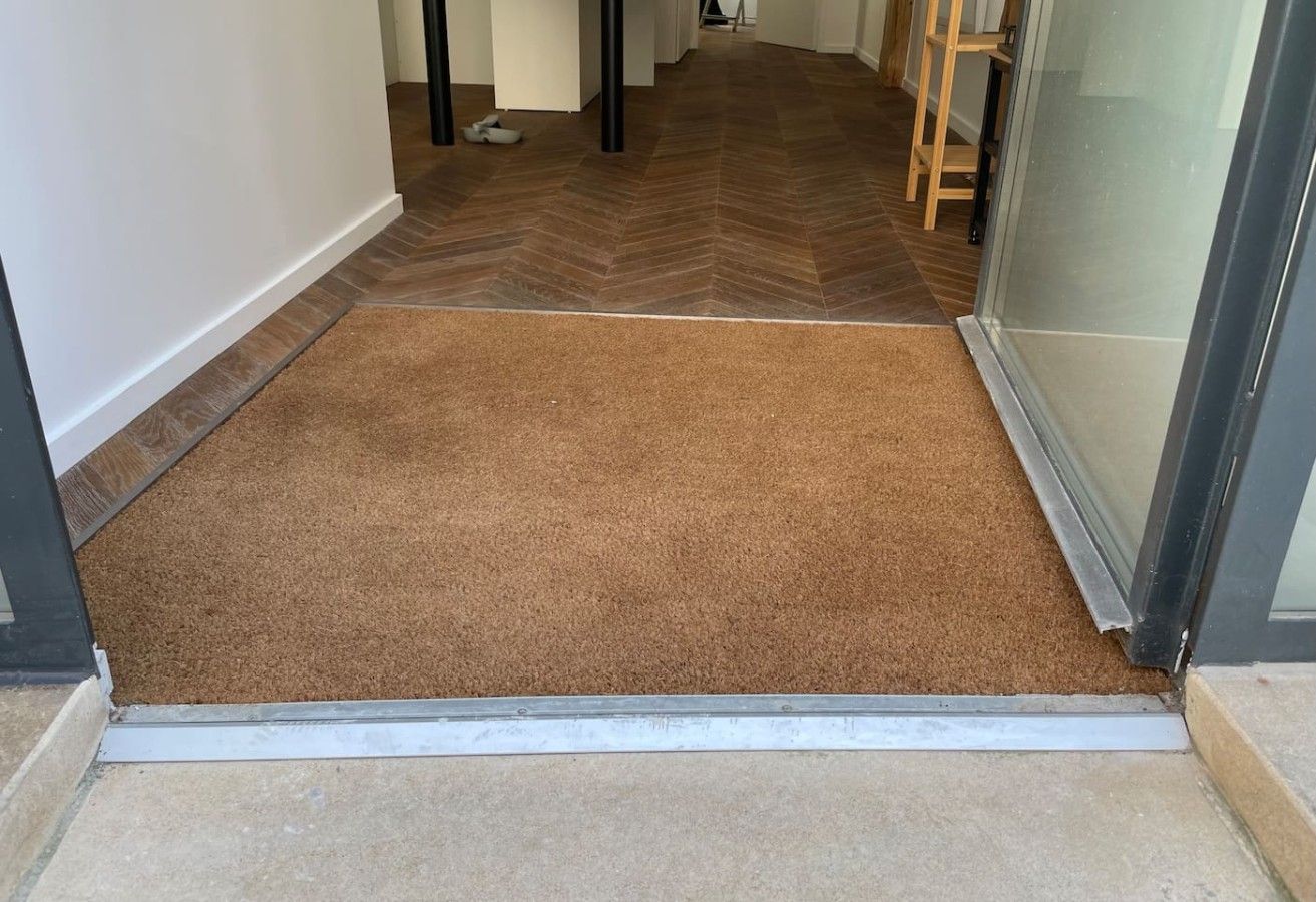 Un couloir avec un tapis marron au niveau de l’entrée, qui mène à une pièce dotée d’une porte en verre.