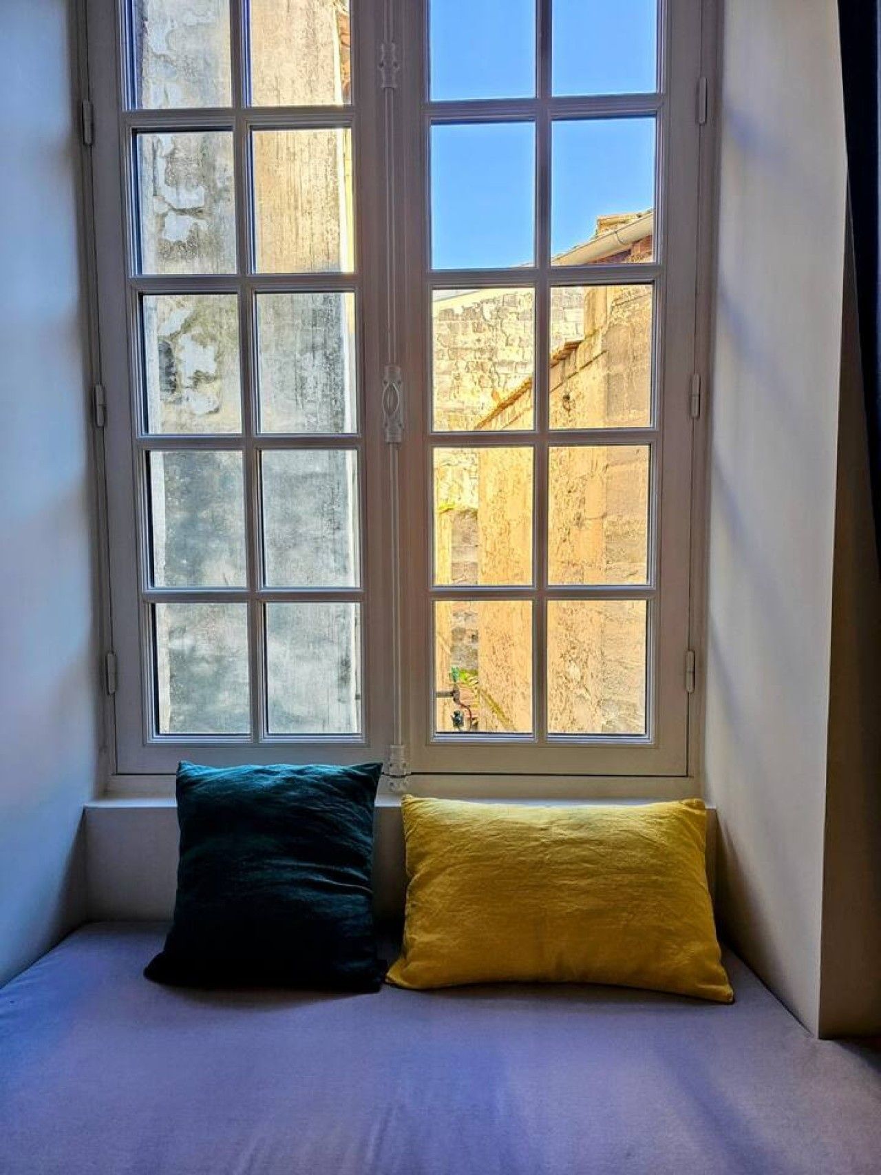 Ein Zimmer mit einem Bett an der Wand, einem Fenster mit Blick auf alte Gebäude und einem Kissen auf dem Bett.