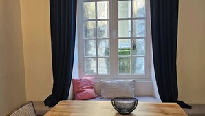 Ein Essbereich mit einem Holztisch und zwei Stühlen, einem Fenster mit schwarzen Vorhängen und einer Bank mit zwei Kissen.
