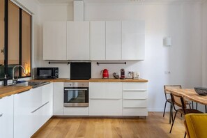 A modern kitchen with white cabinets, a microwave, a stove, and a dining area with a wooden table and chairs. - Chemin du muguet T2 hyper centre (Bordeaux)