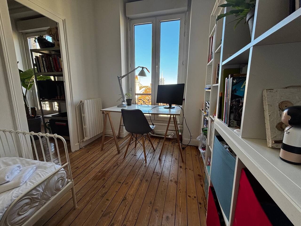 A room with a wooden floor, a desk with a computer, a chair, a radiator, and a bookshelf.
