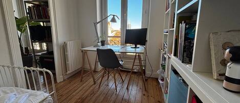 A room with a wooden floor, a desk with a computer, a chair, a radiator, and a bookshelf.