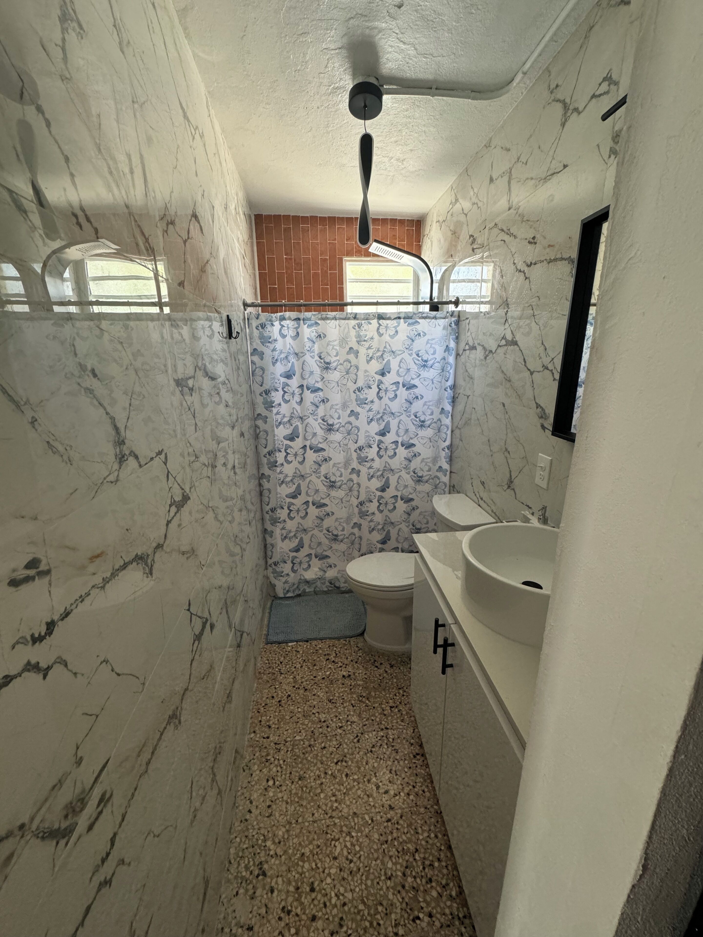 Un baño con paredes de mármol, inodoro, lavabo y cortina de ducha con estampado de mariposas.