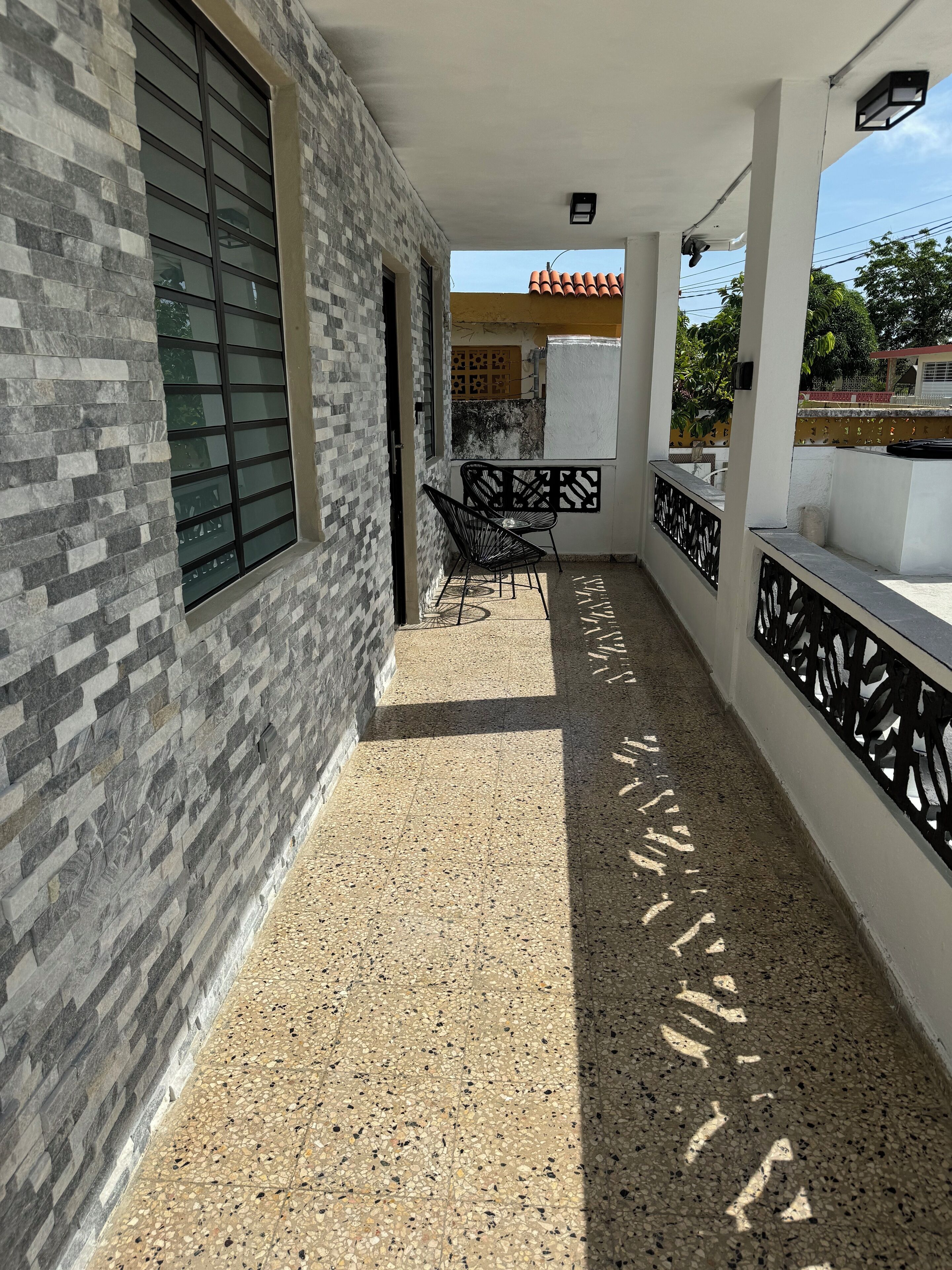 A covered outdoor area with a tiled floor, a seating area with chairs, and a view of a building with a tiled roof.
