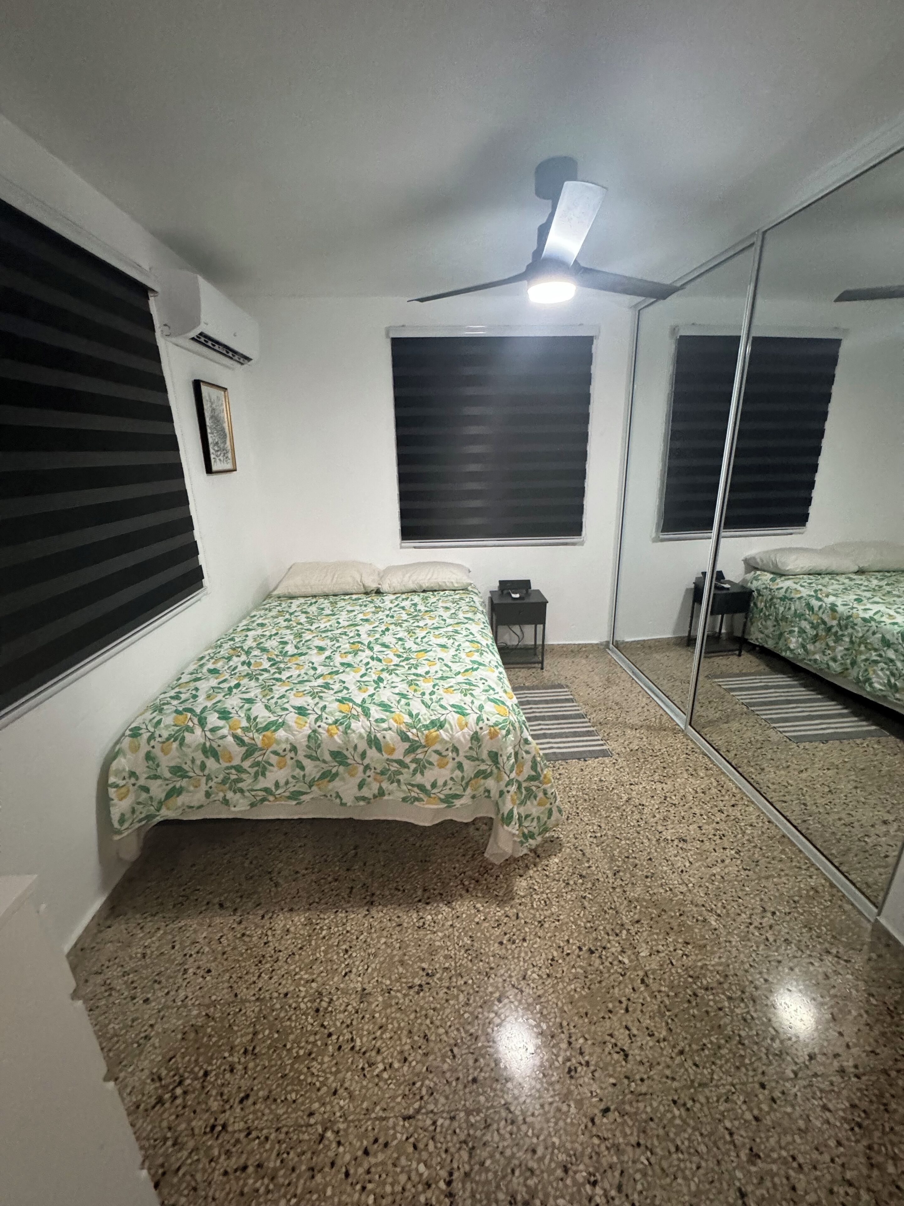 Une chambre à coucher avec un lit, un ventilateur de plafond, une fenêtre avec des stores, un miroir et un sol recouvert de moquette.
