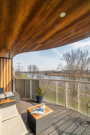 Ein überdachter Außenbereich mit Korbmöbeln, einem Blumentopf und Blick auf einen Fluss und Gebäude in der Ferne.