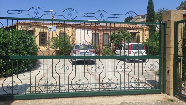 Una entrada tancada amb dues cotxelles aparcades, un edifici amb teulada de rajola i un cel blau clar.