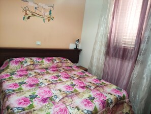 Un dormitorio con una colcha estampada, cabecero de madera, una lámpara de pared y cortinas traslúcidas.