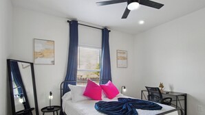 Un dormitorio con cama, escritorio, silla, ventilador de techo y ventana con cortinas.