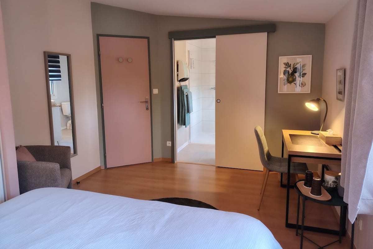 A hotel room with a bed, a desk with a chair, a mirror, and a door leading to a bathroom.