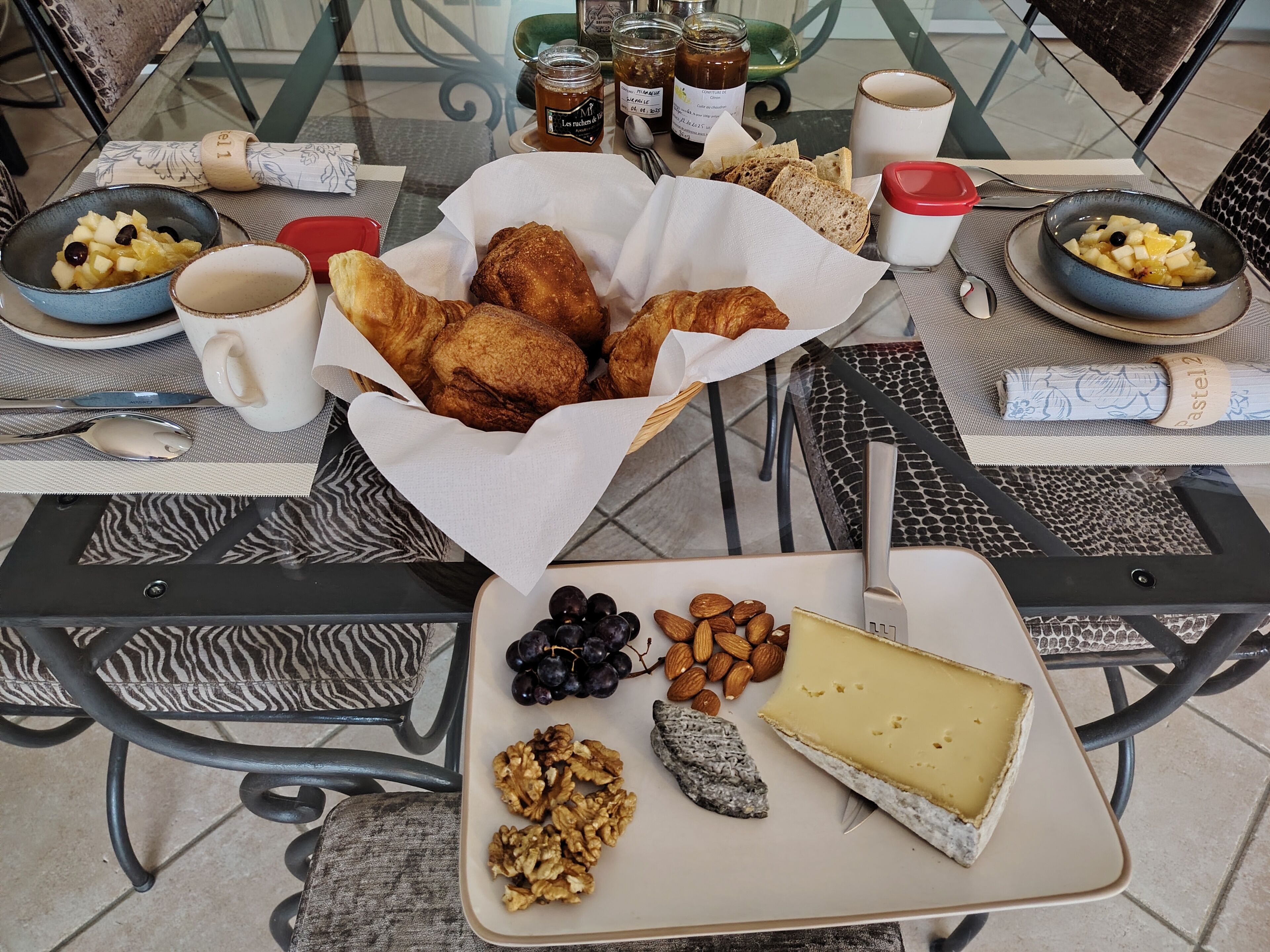 A table with a variety of food items including bread, cheese, grapes, almonds, walnuts, and a jar of jam.