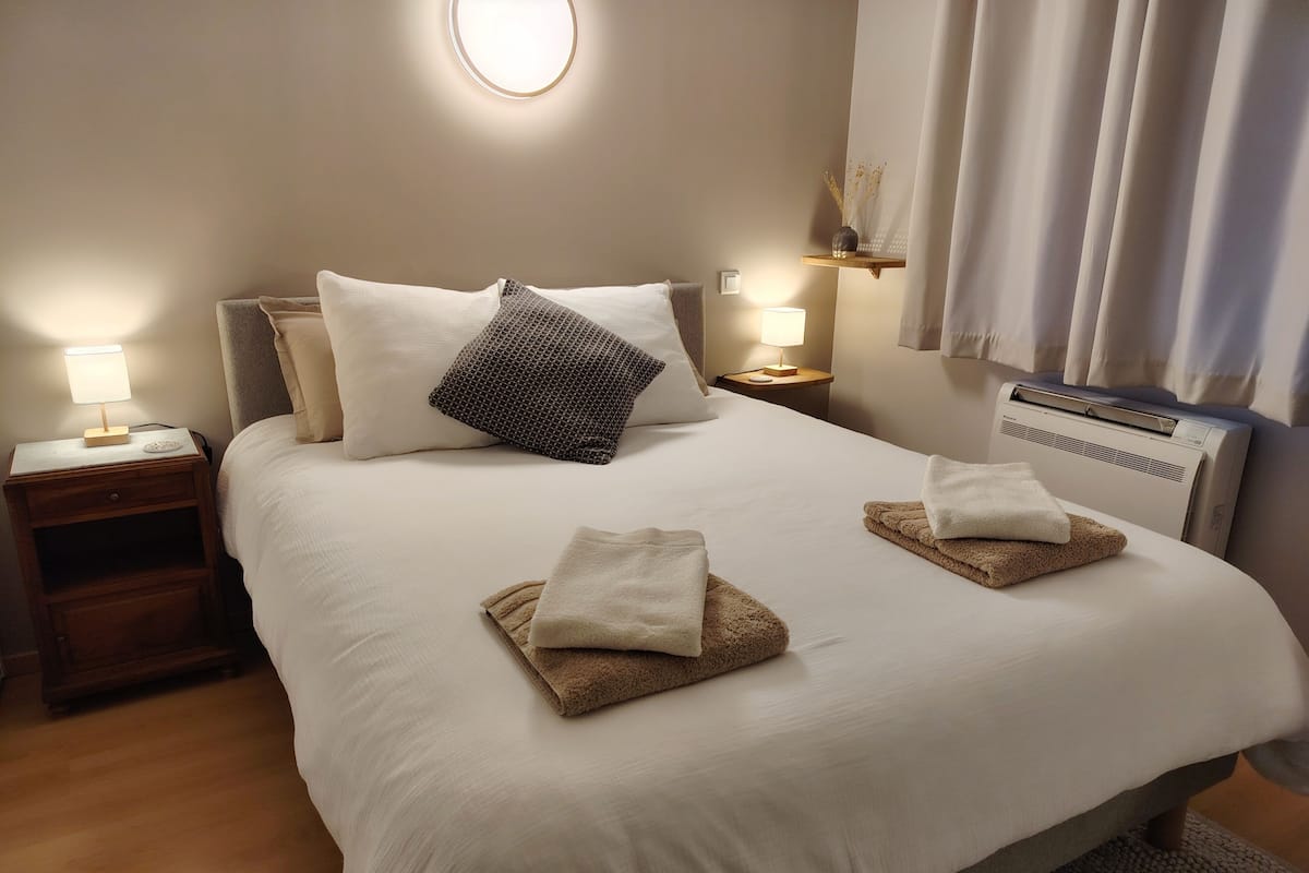 A neatly made bed with two towels, a bedside lamp, a nightstand, and a wall-mounted air conditioner.