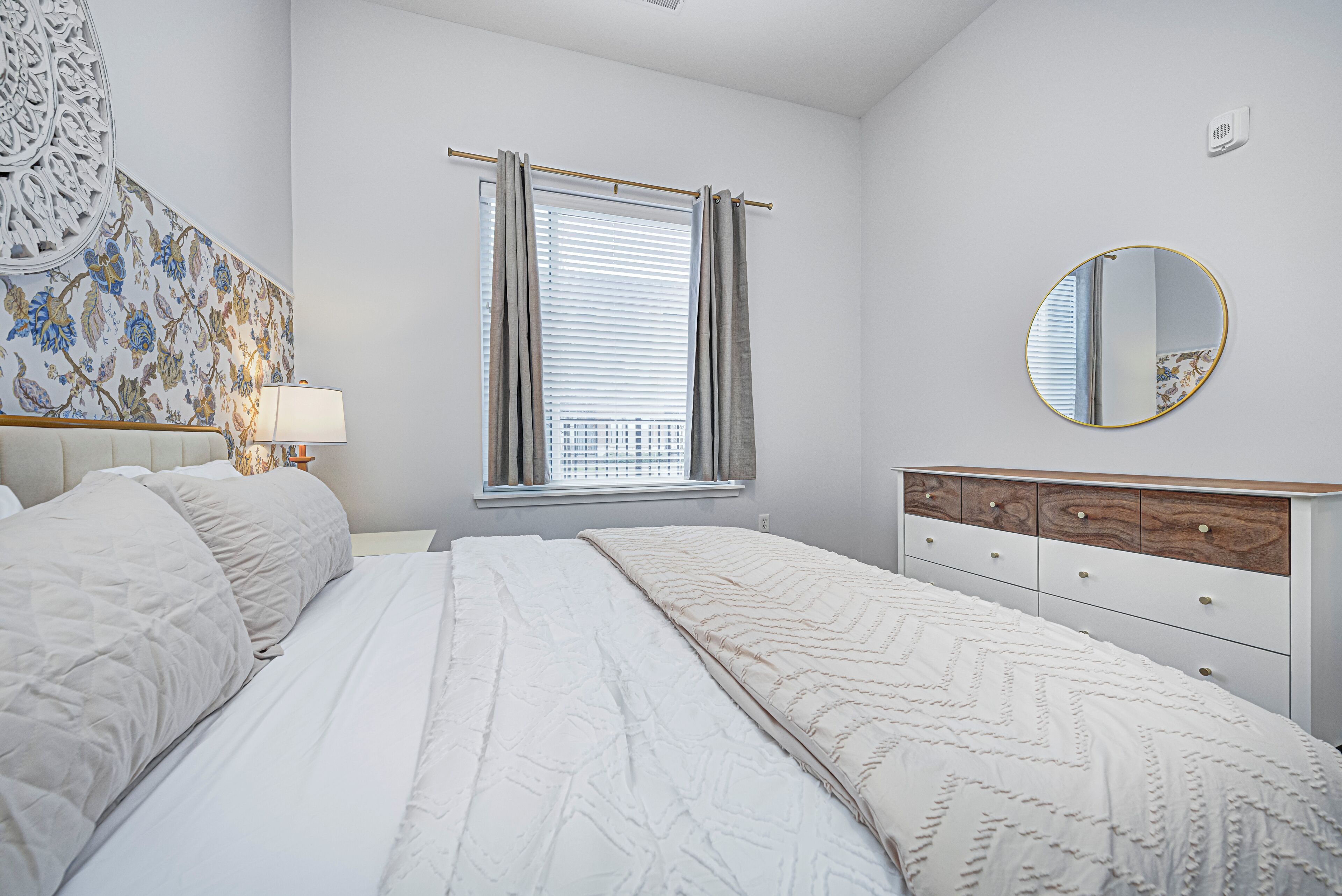 A bedroom with a bed, a bedside table, a lamp, a window with curtains, a mirror, and a wall with a floral pattern.