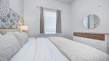 A bedroom with a bed, a bedside table, a lamp, a window with curtains, a mirror, and a wall with a floral pattern.
