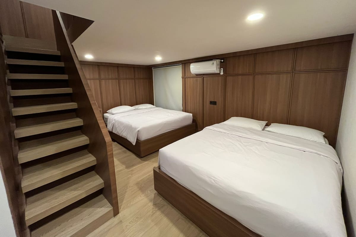 A bedroom with a wooden staircase, two beds, a ceiling-mounted air conditioner, and recessed lighting.
