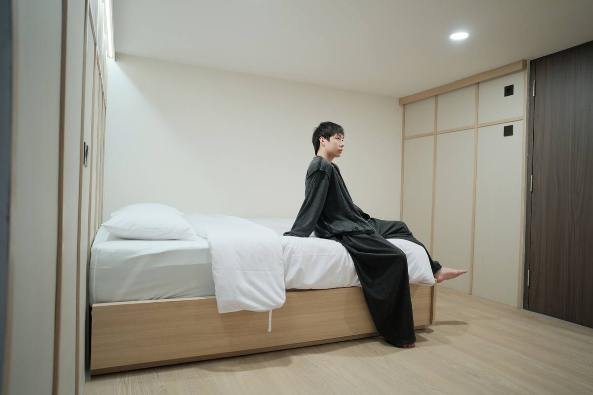 A person sitting on a bed in a room with a wooden floor, a white bedspread, and a wardrobe with sliding doors.