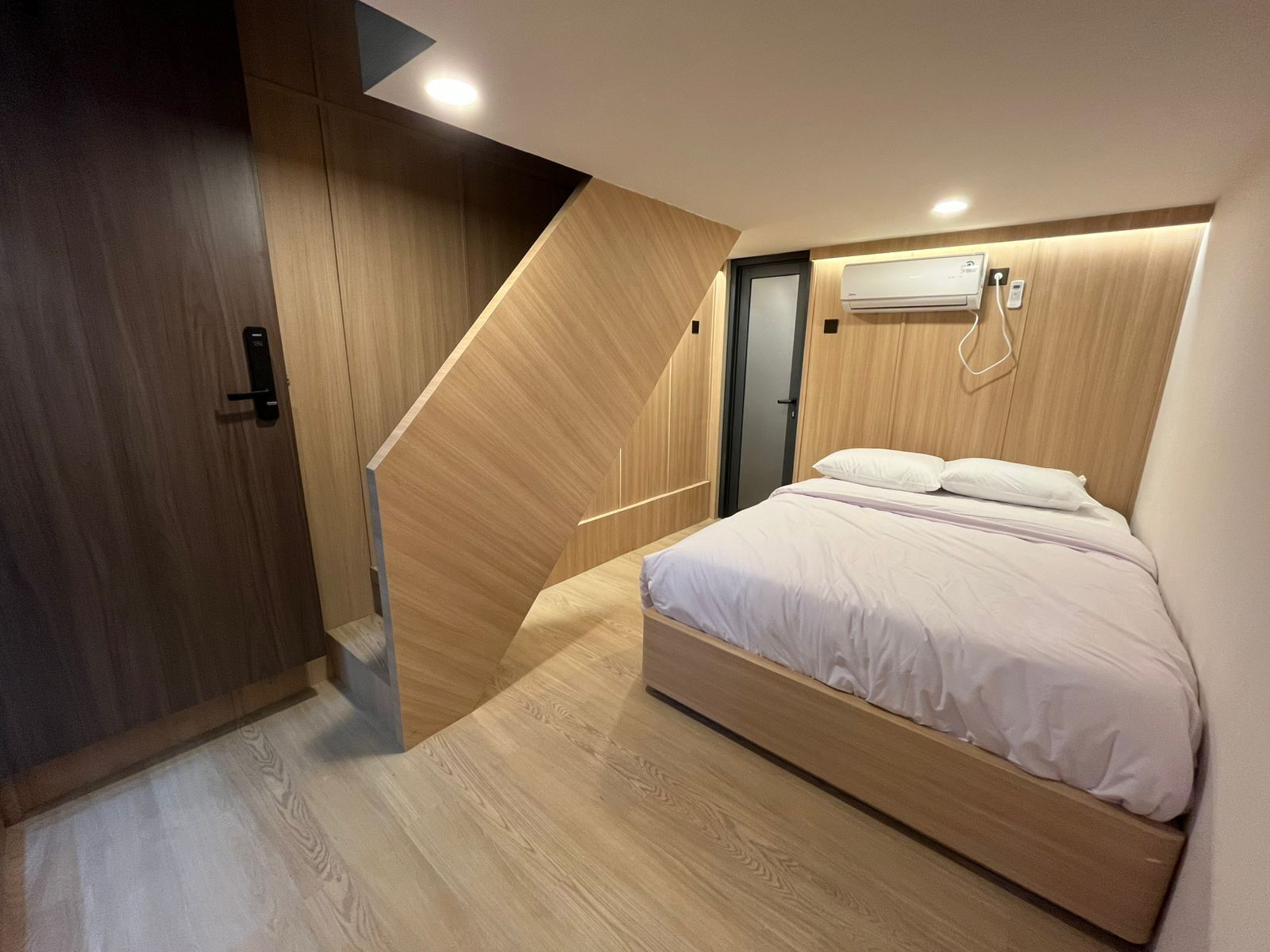 A modern bedroom with a wooden floor, a bed with white bedding, and a built-in air conditioning unit.