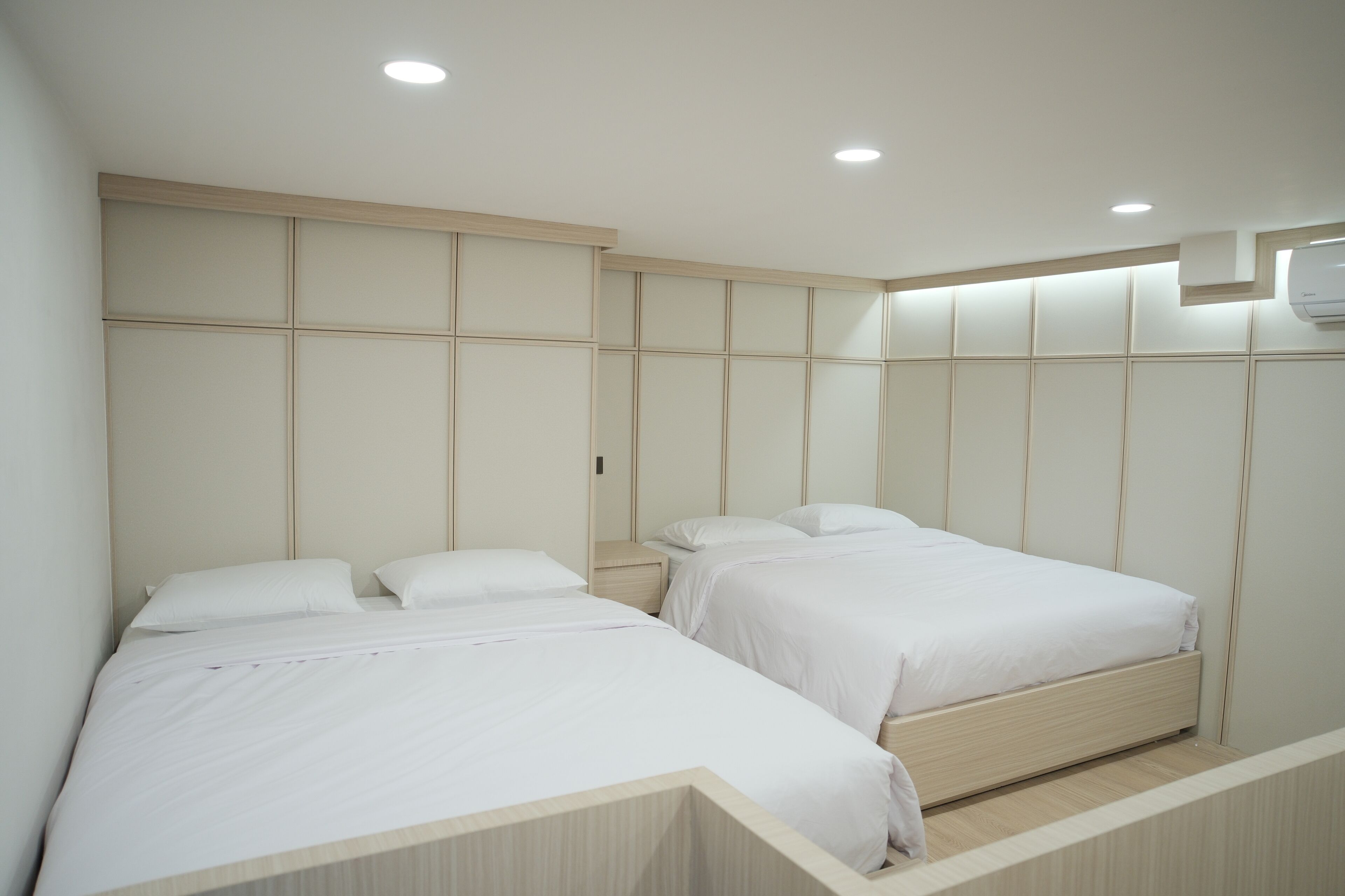 A modern bedroom with two beds, a wooden headboard, and recessed ceiling lights.