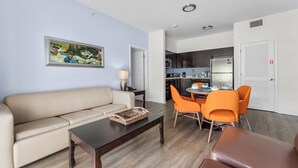 Una sala de estar con un sofá beige, una mesa de centro y sillas naranjas. En el fondo, se aprecia una cocina con armarios oscuros y un refrigerador.