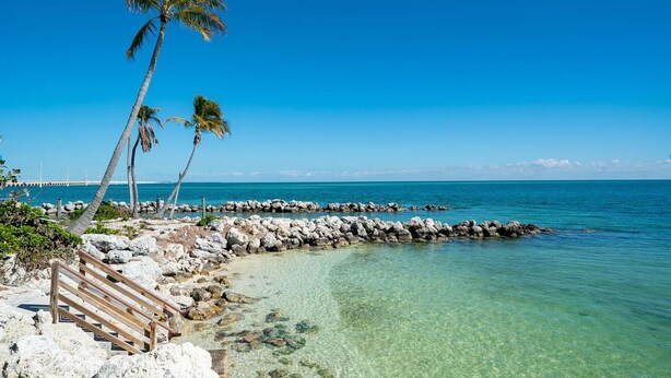 Una playa con agua turquesa cristalina, costa rocosa, palmeras y un paseo marítimo de madera.