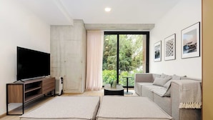 A modern living room with a flat-screen TV, a grey sofa, and a large window with a view of greenery. - w* | Sophisticated 3BR in the Heart of Cuauhtemoc (Ciudad de México)