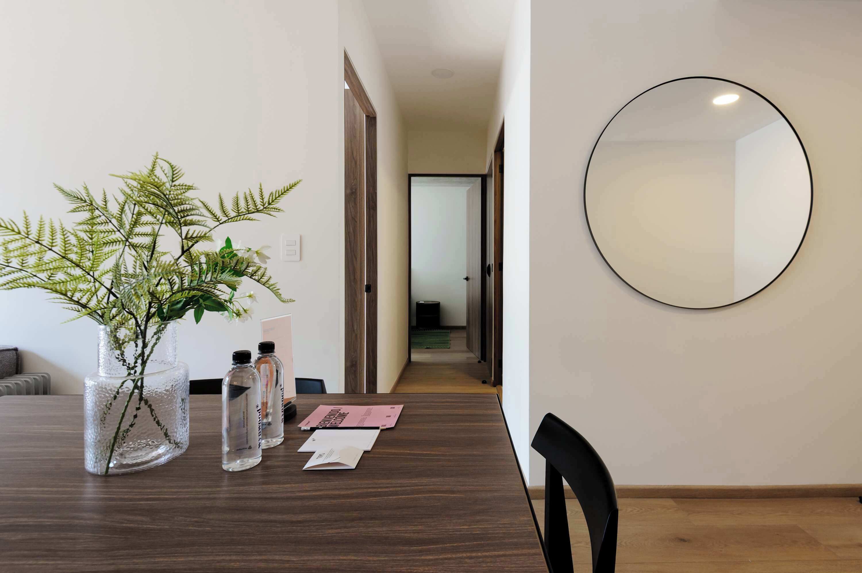 A modern hotel room with a wooden dining table, a vase with green plants, and a round mirror on the wall.