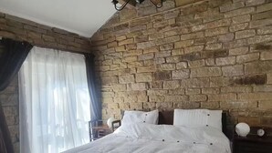 Une chambre avec un mur en pierre, un lit avec une literie blanche, une table de chevet avec une horloge et une fenêtre avec des rideaux légers.