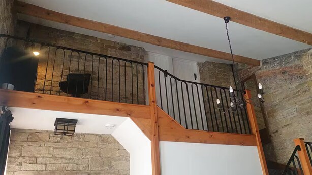 Un intérieur à plusieurs niveaux, avec des poutres en bois apparentes, des murs en pierre et une rampe d’escalier en métal noir.