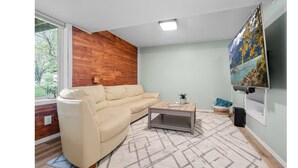 Sala de estar com sofá bege, mesa de centro, TV fixada na parede e janela com vista para árvores.