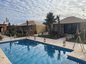 A pool area with lounge chairs and umbrellas, surrounded by palm trees and desert plants. - Agafay Ecolodge (Agafay)