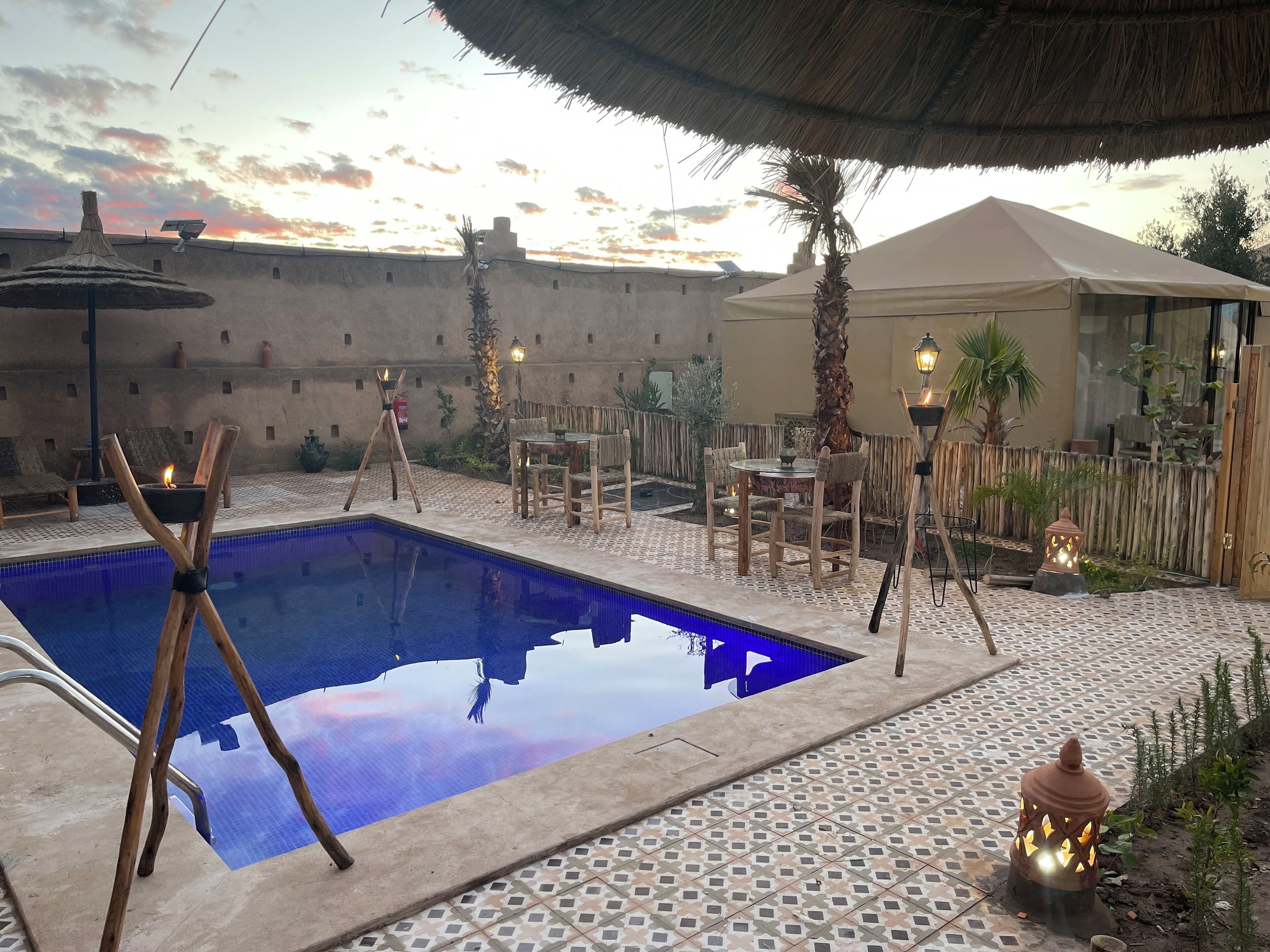 Zona de piscina con piscina azul cristalina, sillas de madera, una mesa y un paraguas de paja.