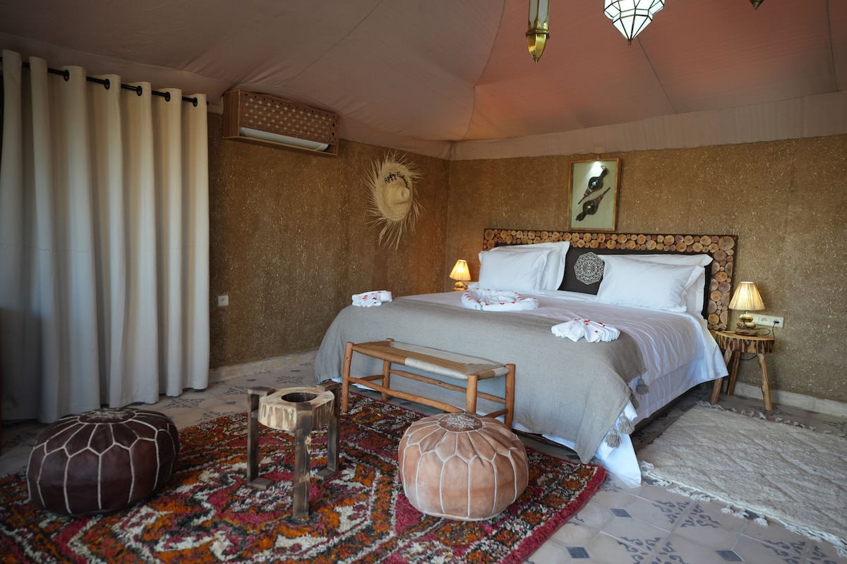 Ein Schlafzimmer mit einem Bett, Nachttischlampen, einem hölzernen Kopfteil, einem gemusterten Teppich und zwei Poufs.