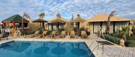 Un área de piscina con sillones y sombrillas, rodeada de palmeras y un paisaje desértico.