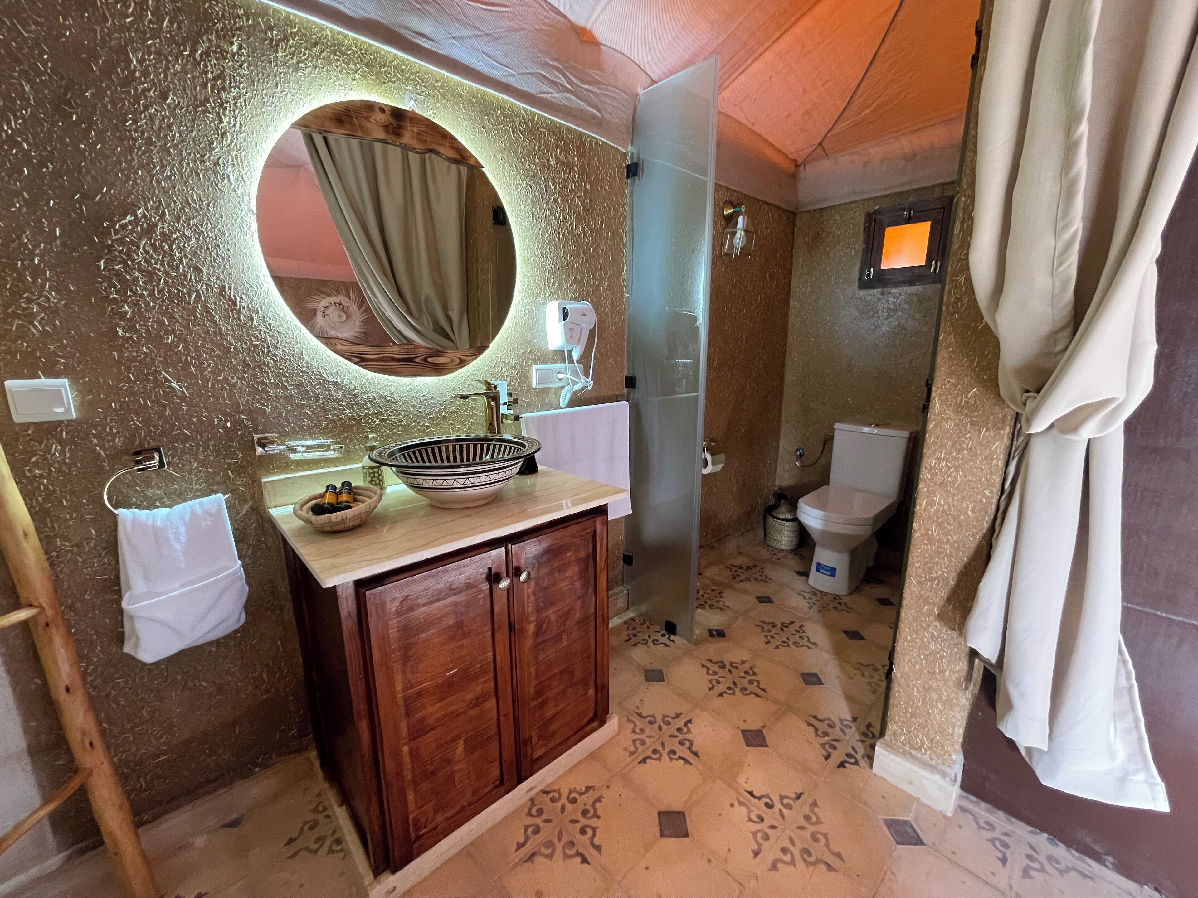 Un baño con tocador de madera, espejo redondo, lavamanos y retrete.