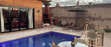A pool area with lounge chairs and a glass table, surrounded by a wooden fence and a brick wall.