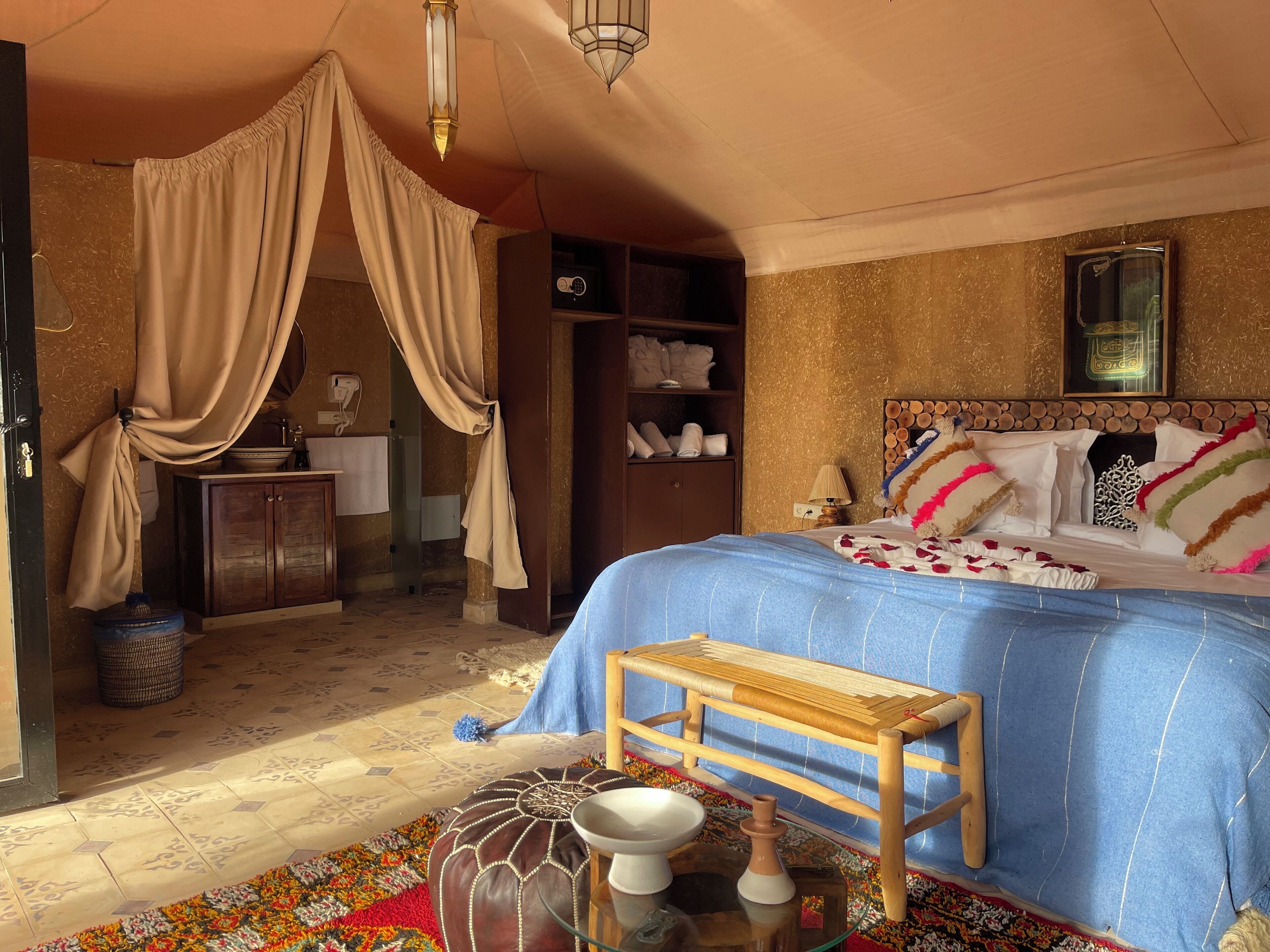 A room with a bed, a wooden bench, a small table, a wall-mounted shelf, and a hanging light fixture.
