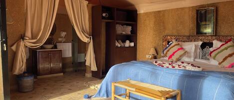 A room with a bed, a wooden bench, a small table, a wall-mounted shelf, and a hanging light fixture.