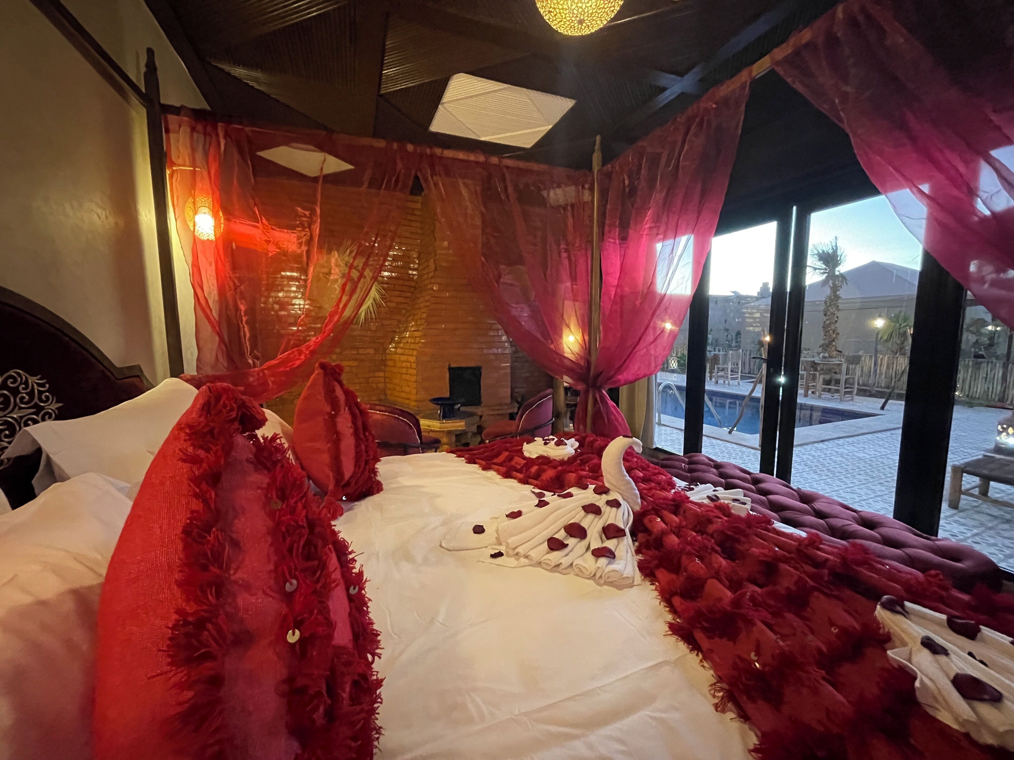 A room with a bed, red curtains, and a view of a pool and palm trees.