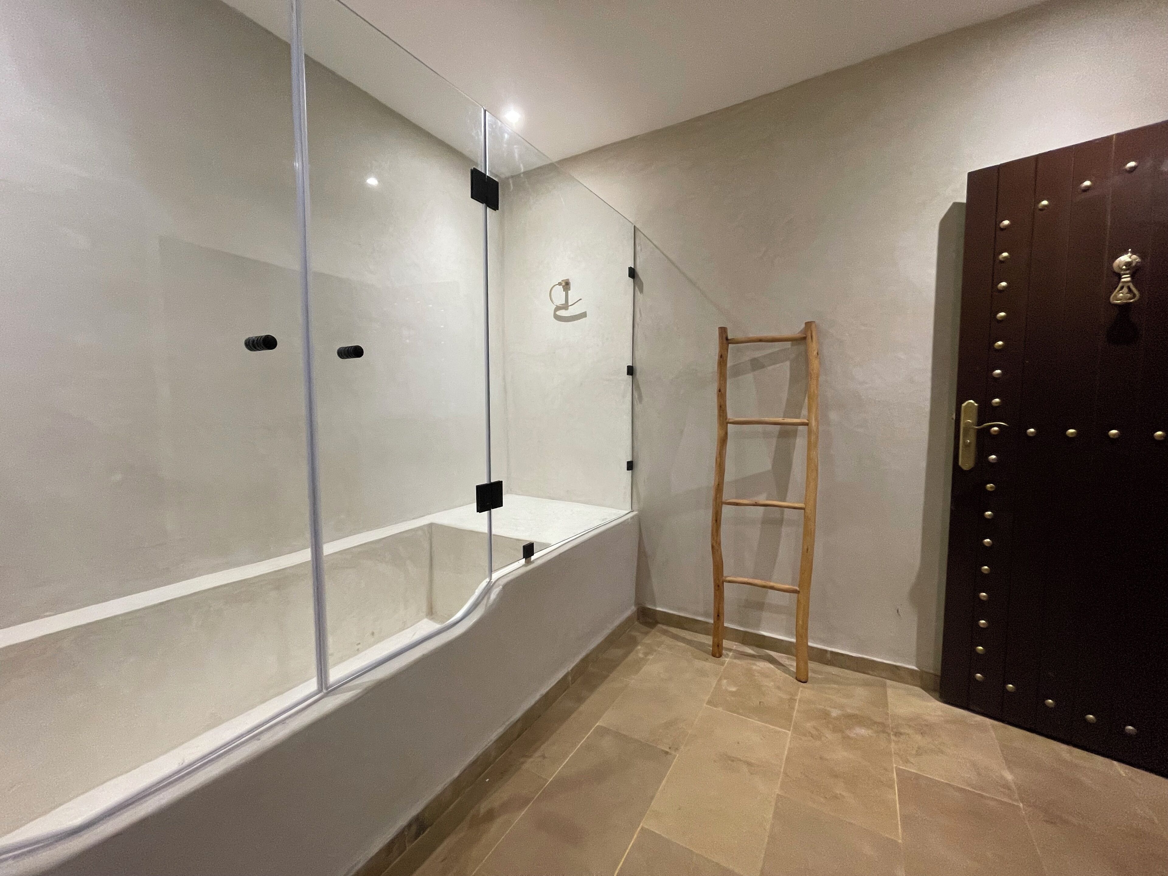 A bathroom with a glass shower enclosure, a wooden ladder, and a dark wooden door.