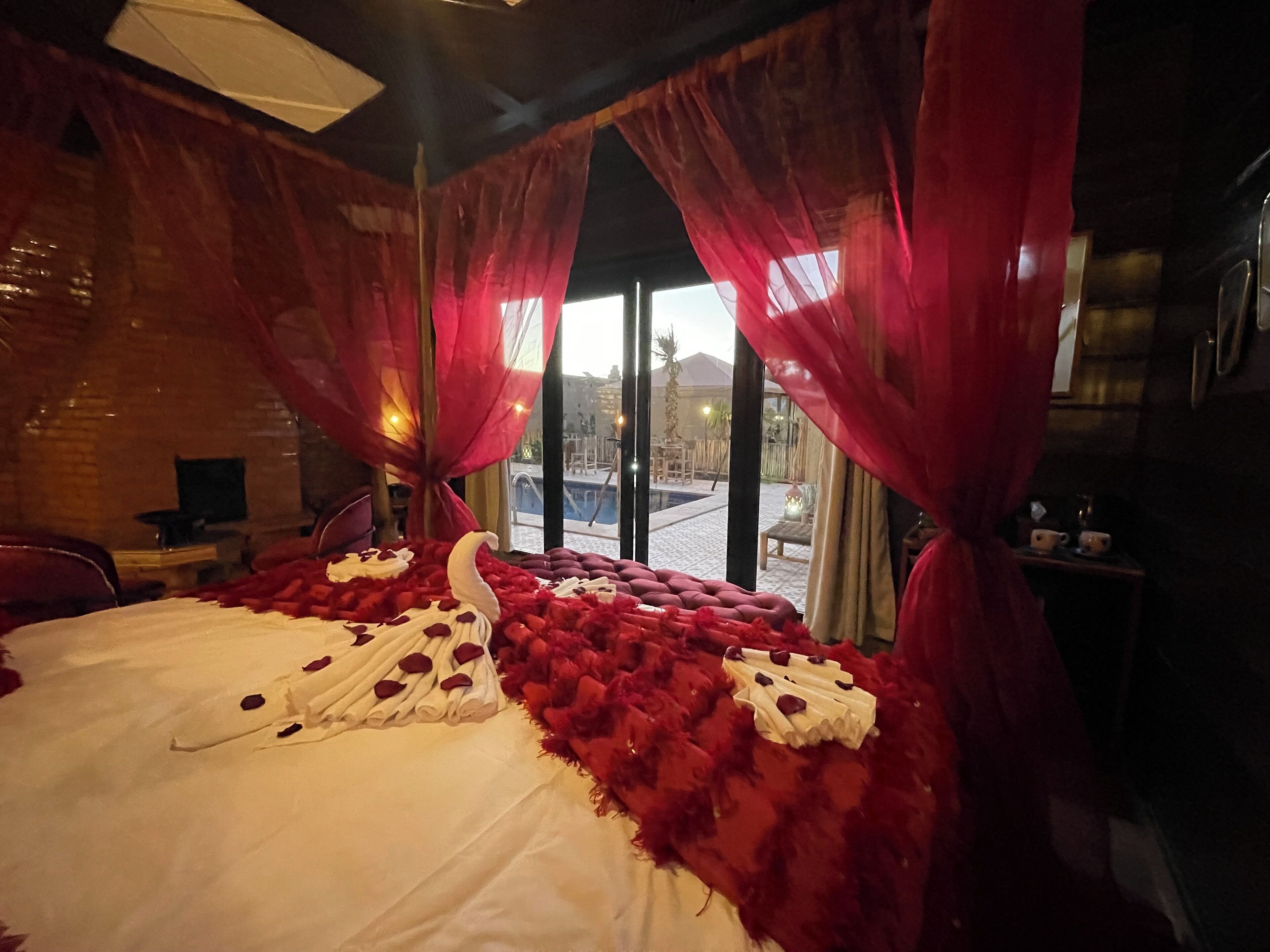 A bed with rose petals and a swan decoration, red curtains, and a view of a pool and buildings outside.