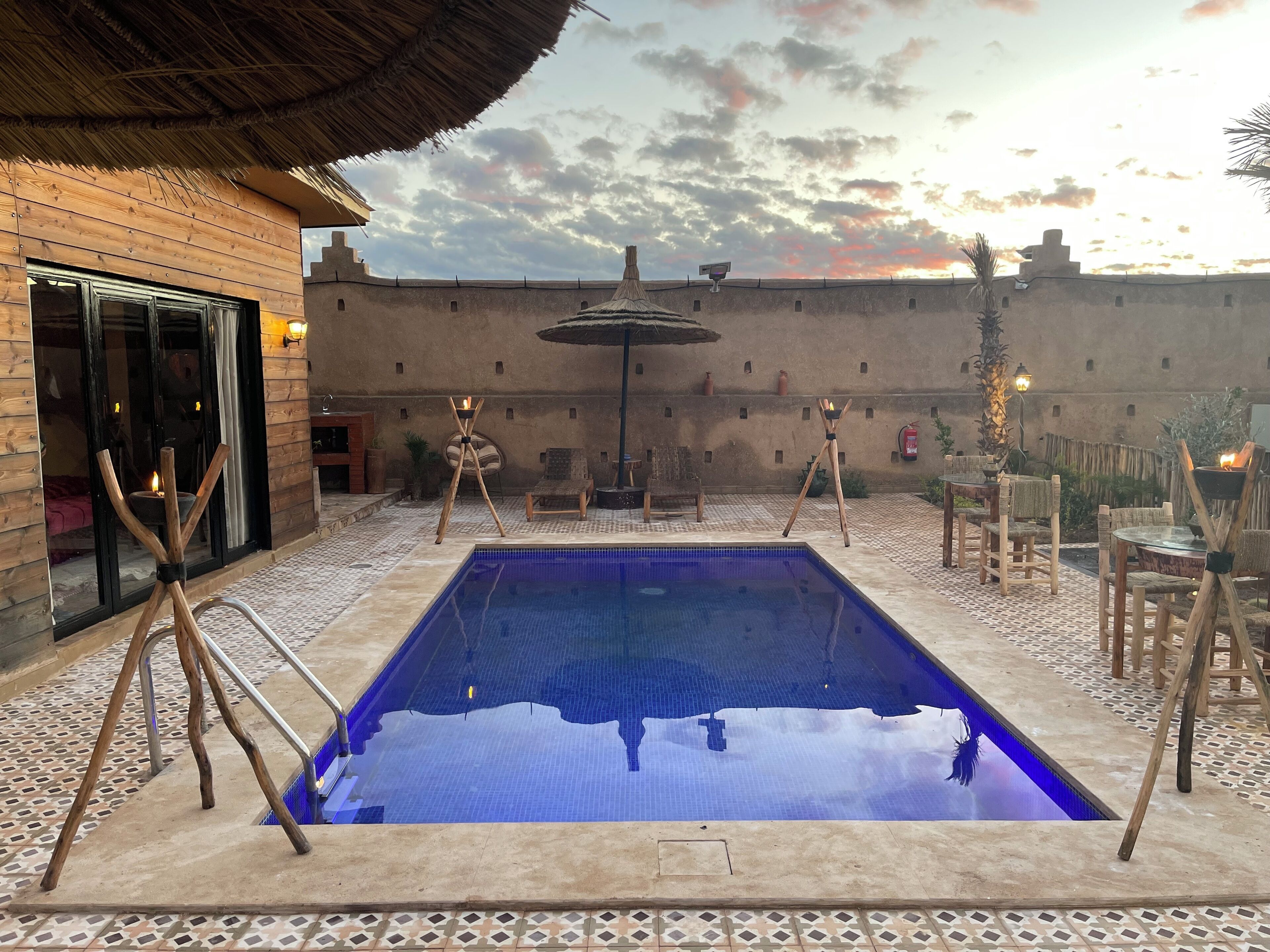 A pool area with a tiled floor, lounge chairs, and a building with wooden walls and a thatched roof.